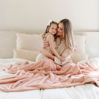 A mother hugs her daughter. They are on a bed under a Blossom (light peach/pink) Lush XL Saranoni blanket, designed for oversized comfort and featuring a buttery soft, plush texture. This luxurious Saranoni blanket is perfect for cozy evenings, reading, or home decor. The Saranoni blanket adds warmth, style, and unmatched softness, making it an ideal gift or essential for ultimate relaxation.