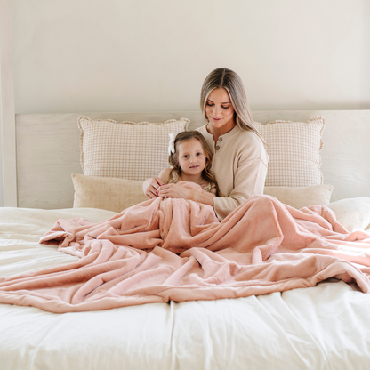 A mother and her daughter sit on a bed under a Blossom (light peach/pink) Lush XL Saranoni blanket, designed for oversized comfort and featuring a buttery soft, plush texture. This luxurious Saranoni blanket is perfect for cozy evenings, reading, or home decor. The Saranoni blanket adds warmth, style, and unmatched softness, making it an ideal gift or essential for ultimate relaxation.