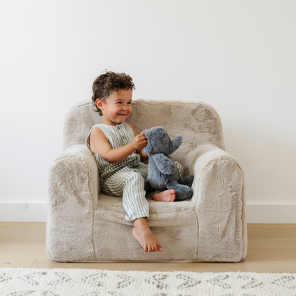 A happy boy sits on a cozy plush chair, joyfully holding a soft gray stuffed elephant. Dressed in a striped outfit, he beams with delight, showcasing the inviting texture of the chair and the comfort of his stuffed animal. This charming scene highlights the perfect blend of stylish children's furniture and beloved stuffed animals, making it an ideal addition to any child's playroom or reading nook.