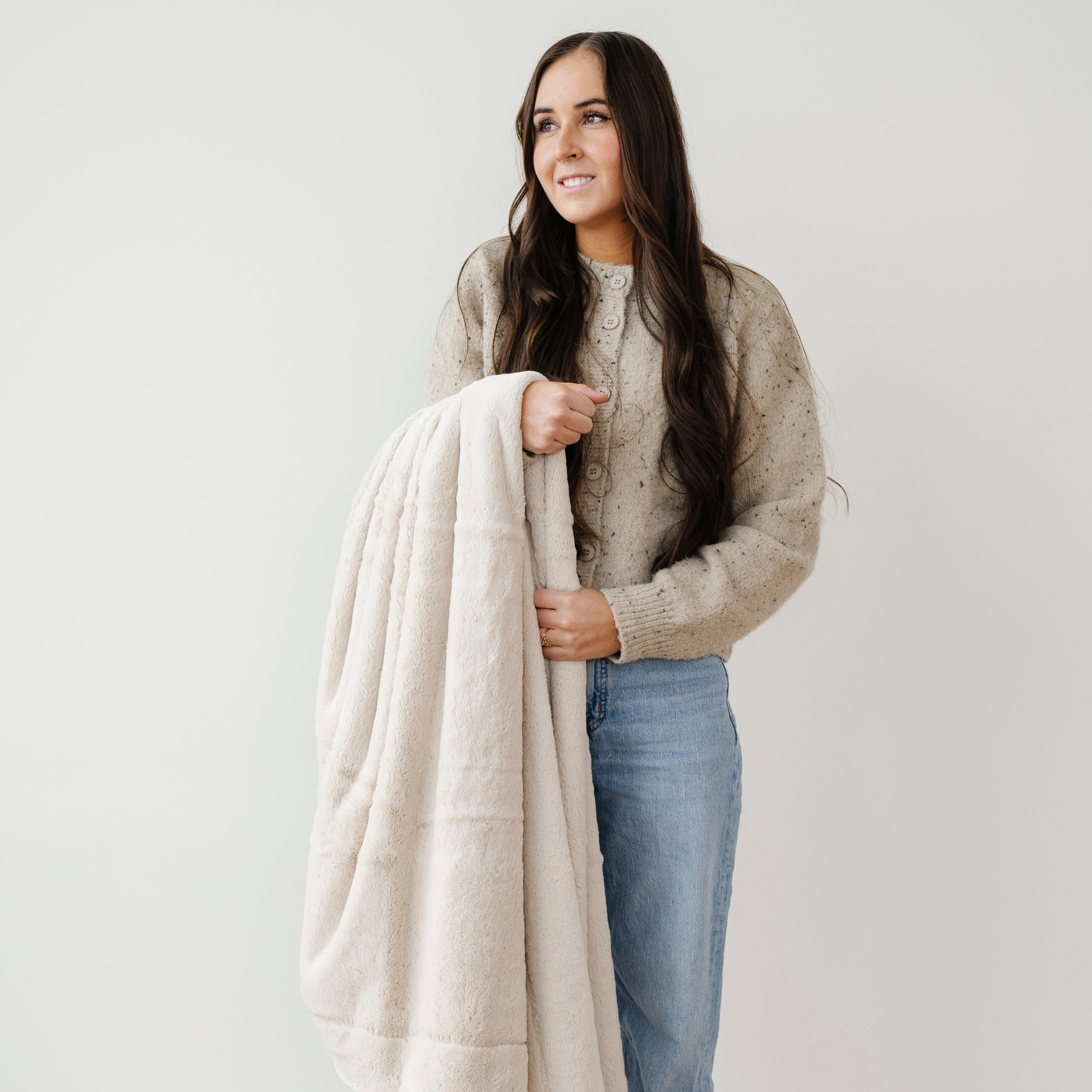A woman smiles and looks into the distance as she holds a light tan lush Saranoni blanket. This cozy blanket is a luxury blanket and a faux fur blanket. This soft blanket is great as a home decor staple, comfort gift, or everyday comfort item. Elevate your home and home decor with this faux fur blanket that is also a throw blanket. Enjoy this luxury blanket for years to come.