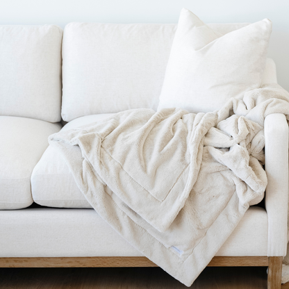 A light tan luxury lush Saranoni blanket is draped on the side of a couch. This cozy blanket is a luxury blanket and a faux fur blanket. This soft blanket is great as a home decor staple, gift, or everyday comfort item. Elevate your home and home decor with this faux fur blanket that is also a throw blanket. Enjoy this luxury blanket for years to come.