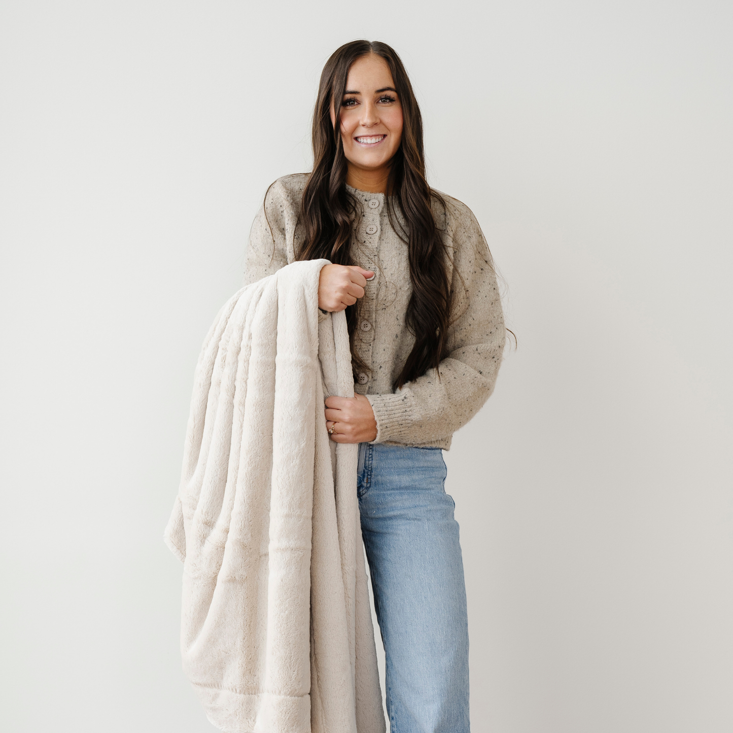 A woman smiles an holds a light tan lush Saranoni blanket. This luxury blanket is a cozy blanket and a faux fur blanket. This soft blanket is great as a home decor staple, comfort gift, or everyday comfort item. Elevate your home and home decor with this faux fur blanket that is also a throw blanket. Enjoy this cozy blanket for years to come.