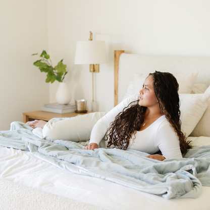 A woman lays on a bed on a Chambray (light blue) Lush XL Saranoni blanket, designed for oversized comfort and featuring a buttery soft, plush texture. This luxurious Saranoni blanket is perfect for cozy evenings, reading, or home decor. The Saranoni blanket adds warmth, style, and unmatched softness, making it an ideal gift or essential for ultimate relaxation.