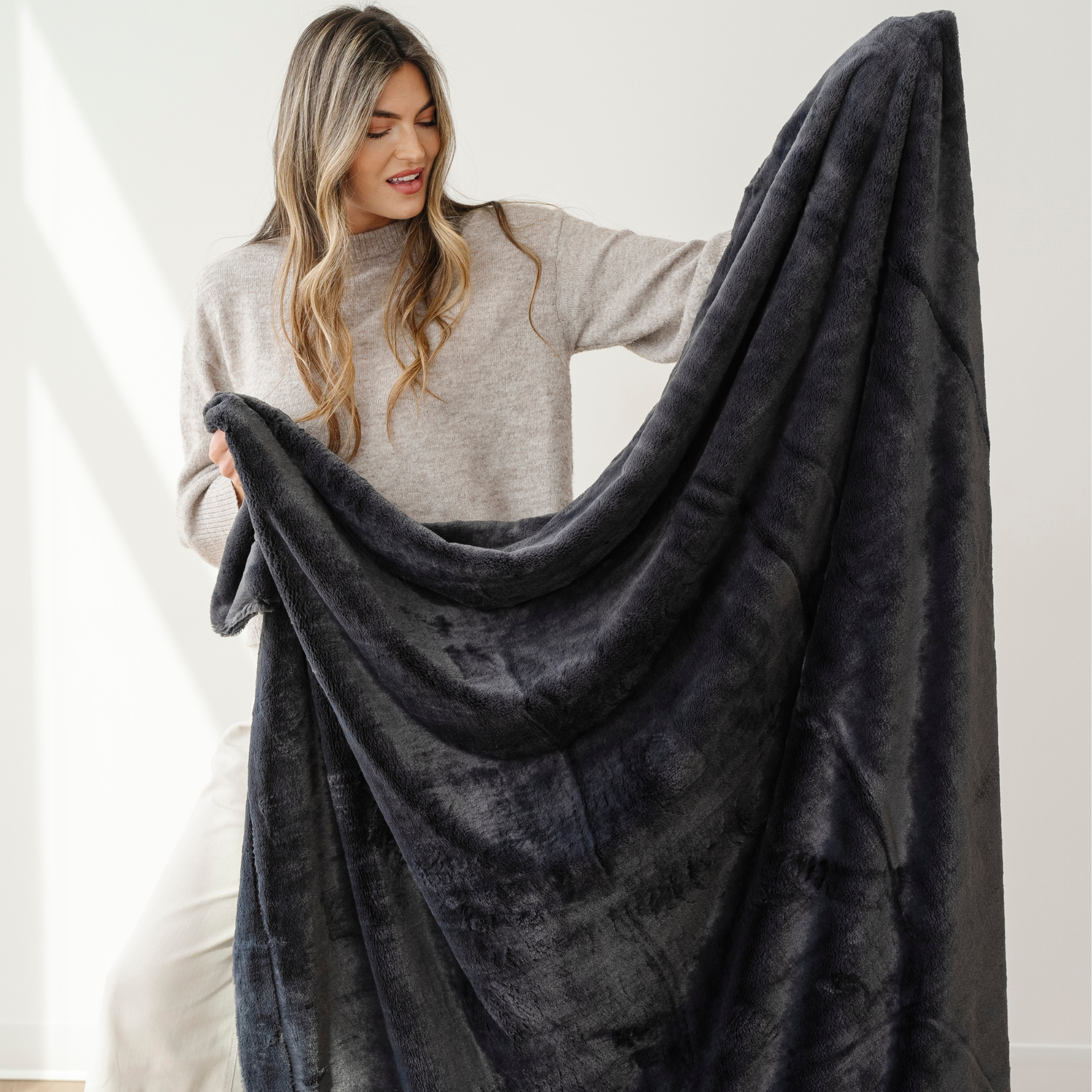 A woman holds up a charcoal lush Saranoni blanket. This luxury blanket is a cozy blanket and a faux fur blanket. This soft blanket is great as a home decor staple, comfort gift, or everyday comfort item. Elevate your home and home decor with this faux fur blanket that is also a throw blanket. Enjoy this soft throw blanket for years to come.