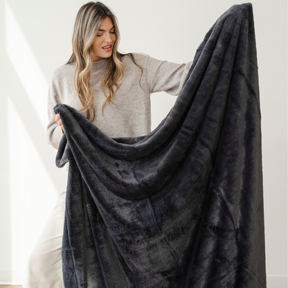 A woman holds up a charcoal lush Saranoni blanket. This luxury blanket is a cozy blanket and a faux fur blanket. This soft blanket is great as a home decor staple, comfort gift, or everyday comfort item. Elevate your home and home decor with this faux fur blanket that is also a throw blanket. Enjoy this soft throw blanket for years to come.