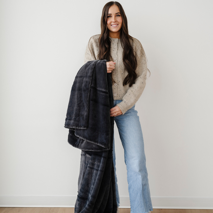 A woman smiles and holds a charcoal blanket that is a lush blanket with a luxurious texture. This cozy blanket is great for gifting, relaxing, or as an addition to your home decor. This soft blanket is a luxury blanket and a Saranoni blanket. Enjoy luxury that lasts.