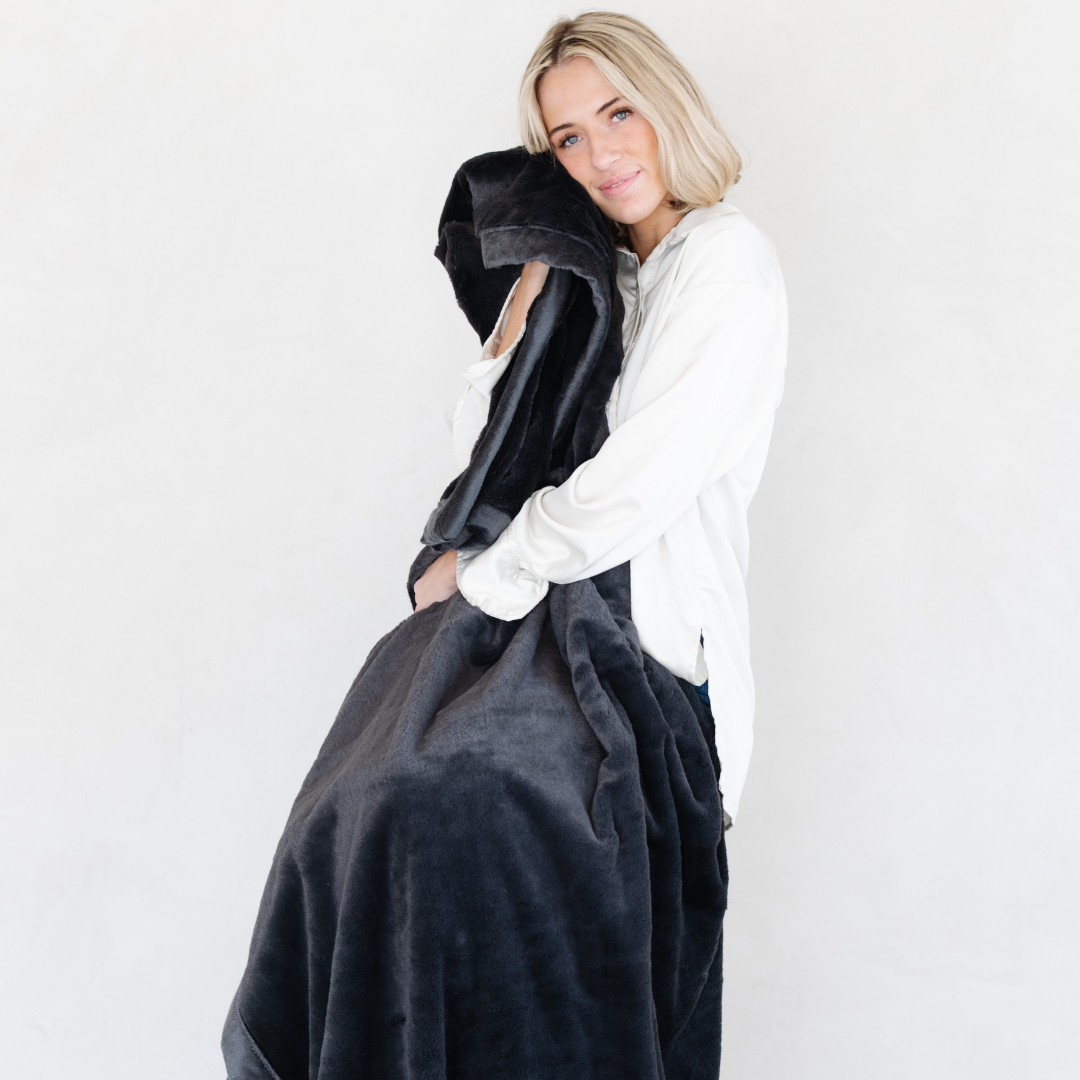 A woman holds a Charcoal Lush XL Saranoni blanket, designed for oversized comfort and featuring a buttery soft, plush texture. This luxurious Saranoni blanket is perfect for cozy evenings, reading, or home decor. The Saranoni blanket adds warmth, style, and unmatched softness, making it an ideal gift or essential for ultimate relaxation.