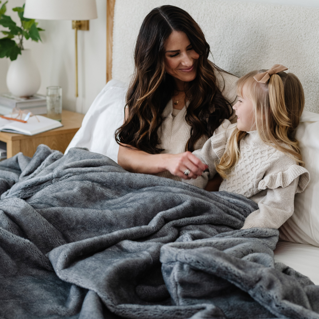 A mother and her daughter smile and sit under a Saranoni Grand Faux Fur blanket, featuring an elegant light charcoal tone and luxuriously soft texture. This cozy, queen-sized blanket and faux fur blanket offers soothing weight, premium softness, timeless comfort, and is a soft blanket. Perfect as a big blanket and luxury blanket for snuggling or as a luxury bedroom accent, this Saranoni blanket elevates every cozy moment and is a cozy blanket.