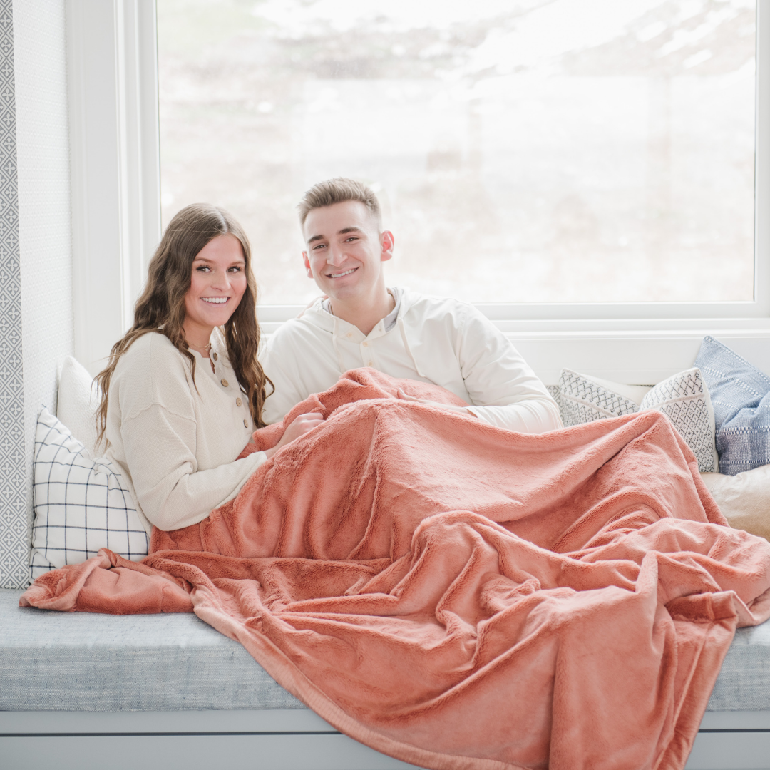 A husband and wife smile and sit under a Clay (peach) Lush XL Saranoni blanket, designed for oversized comfort and featuring a buttery soft, plush texture. This luxurious Saranoni blanket is perfect for cozy evenings, reading, or home decor. The Saranoni blanket adds warmth, style, and unmatched softness, making it an ideal gift or essential for ultimate relaxation.