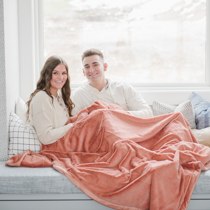 A husband and wife smile and sit under a Clay (peach) Lush XL Saranoni blanket, designed for oversized comfort and featuring a buttery soft, plush texture. This luxurious Saranoni blanket is perfect for cozy evenings, reading, or home decor. The Saranoni blanket adds warmth, style, and unmatched softness, making it an ideal gift or essential for ultimate relaxation.