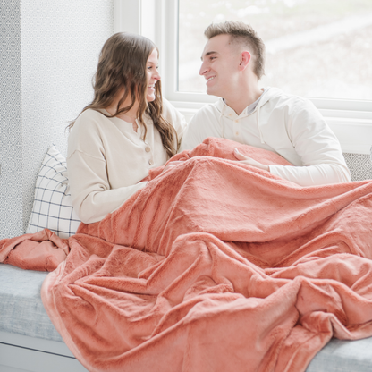 A husband and wife sit under a Clay (peach) Lush XL Saranoni blanket, designed for oversized comfort and featuring a buttery soft, plush texture. This luxurious Saranoni blanket is perfect for cozy evenings, reading, or home decor. The Saranoni blanket adds warmth, style, and unmatched softness, making it an ideal gift or essential for ultimate relaxation.