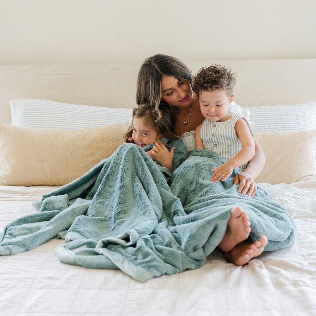 A mother and her son and daughter sit with a Eucalyptus Lush XL Saranoni blanket, designed for oversized comfort and featuring a buttery soft, plush texture, on their laps. This luxurious Saranoni blanket is perfect for cozy evenings, reading, or home decor. The Saranoni blanket adds warmth, style, and unmatched softness, making it an ideal gift or essential for ultimate relaxation.