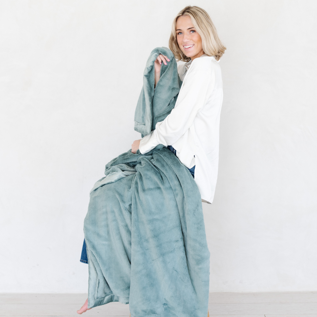 A woman holds a Eucalyptus Lush XL Saranoni blanket, designed for oversized comfort and featuring a buttery soft, plush texture. This luxurious Saranoni blanket is perfect for cozy evenings, reading, or home decor. The Saranoni blanket adds warmth, style, and unmatched softness, making it an ideal gift or essential for ultimate relaxation.