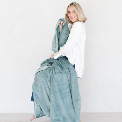 A woman holds a Eucalyptus Lush XL Saranoni blanket, designed for oversized comfort and featuring a buttery soft, plush texture. This luxurious Saranoni blanket is perfect for cozy evenings, reading, or home decor. The Saranoni blanket adds warmth, style, and unmatched softness, making it an ideal gift or essential for ultimate relaxation.