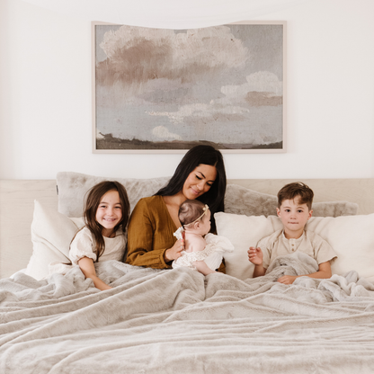 A mother and her children sit under a Saranoni Grand Faux Fur blanket, featuring an elegant light cream/gray tone and luxuriously soft texture. This cozy, queen-sized blanket and faux fur blanket offers soothing weight, premium softness, timeless comfort, and is a soft blanket. Perfect as a big blanket and luxury blanket for snuggling or as a luxury bedroom accent, this Saranoni blanket elevates every cozy moment and is a cozy blanket.