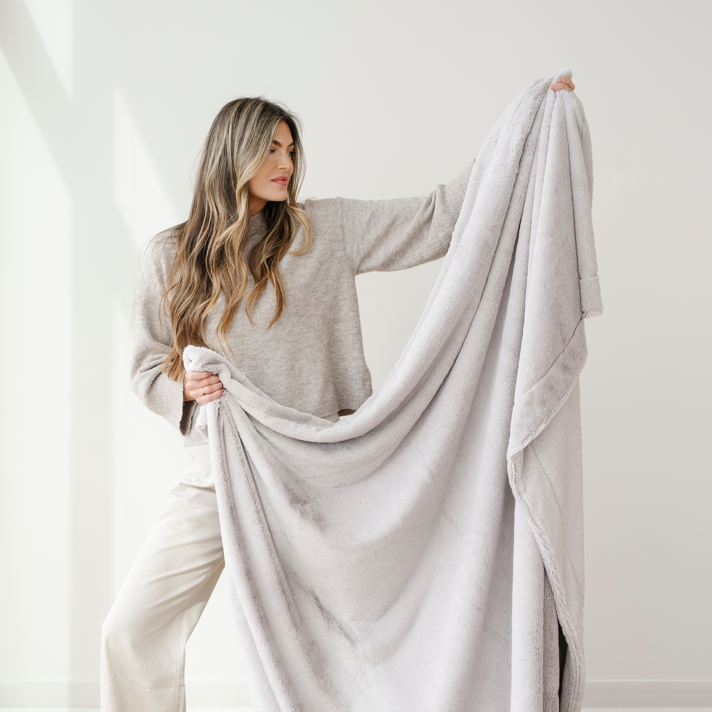 A woman holds up a light gray lush Saranoni blanket. This soft throw blanket is a luxury blanket and a faux fur blanket. This soft blanket is great as a home decor staple, comfort gift, or everyday comfort item. Elevate your home and home decor with this faux fur blanket that is also a throw blanket. Enjoy this cozy blanket for years to come.