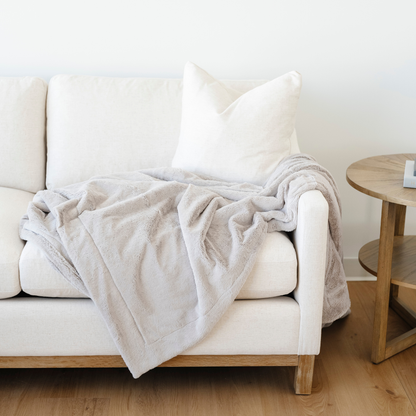 A light gray lush Saranoni blanket that is draped on a couch. This cozy blanket is a luxury blanket and a faux fur blanket. This soft blanket is great as a home decor staple, comfort gift, or everyday comfort item. Elevate your home and home decor with this faux fur blanket that is also a throw blanket. Enjoy this cozy blanket for years to come.