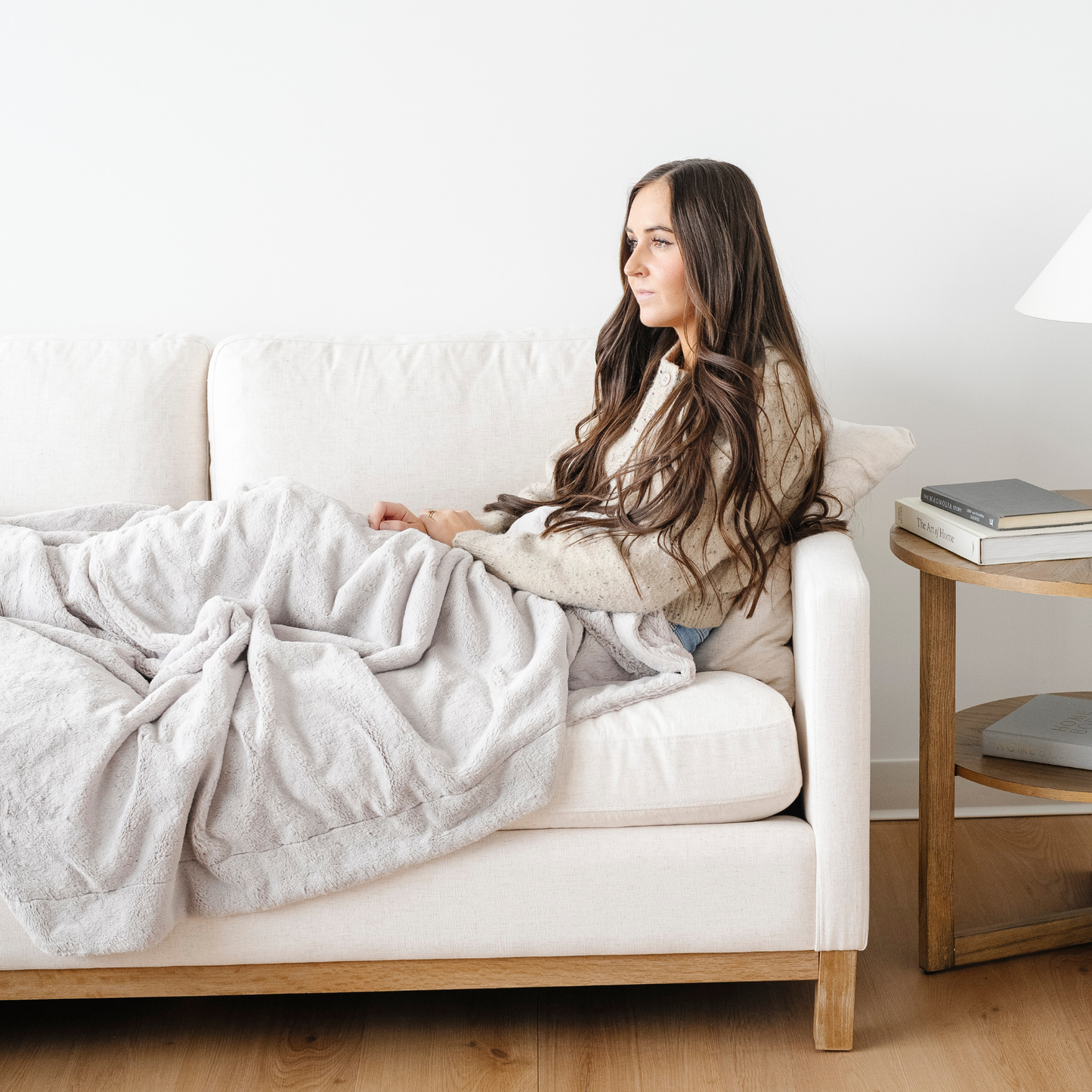 A woman sits on a couch with a light gray lush Saranoni blanket on her lap. This luxury blanket is a soft throw blanket and a faux fur blanket. This soft blanket is great as a home decor staple, comfort gift, or everyday relaxation item. Elevate your home and home decor with this faux fur blanket that is also a throw blanket. Enjoy this cozy blanket for years to come.