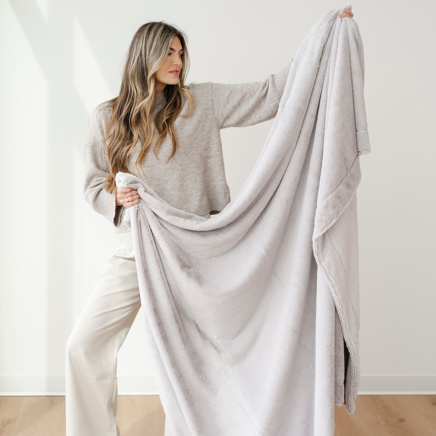 A woman holds up a soft blanket that is a light gray cozy blanket. This luxury blanket is also an extra large blanket and a Saranoni blanket. This faux fur blanket is great for gifting, relaxing, and elevating your home as a home decor staple. 