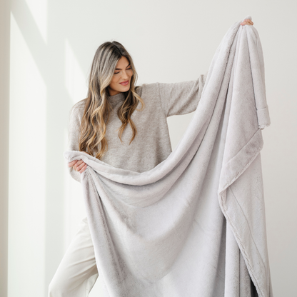 A woman smiles and holds up a light gray lush Saranoni blanket. This luxury blanket is a soft throw blanket and a faux fur blanket. This soft blanket is great as a home decor staple, comfort gift, or everyday comfort item. Elevate your home and home decor with this faux fur blanket that is also a throw blanket. Enjoy this cozy blanket for years to come.