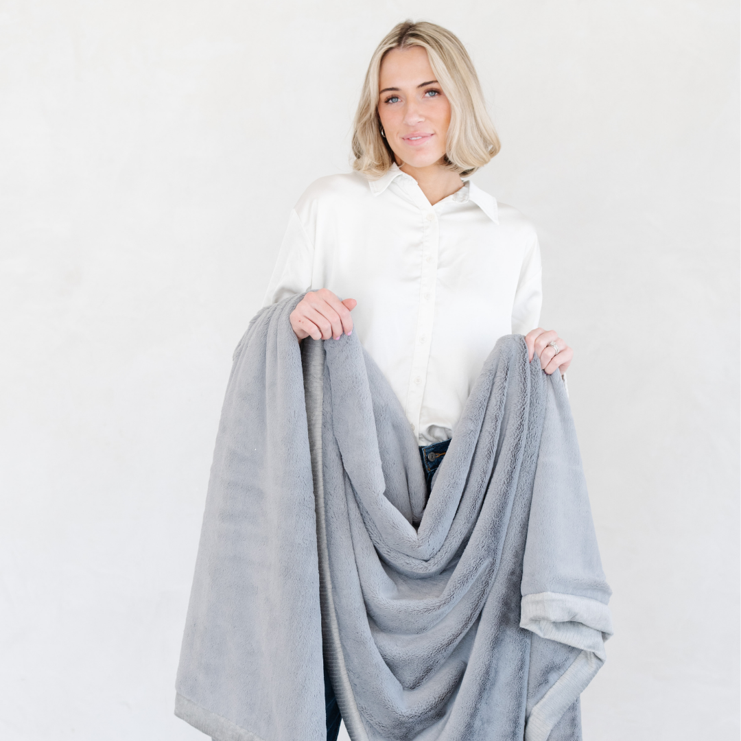 A woman holds a Gray Lush XL Saranoni blanket, designed for oversized comfort and featuring a buttery soft, plush texture. This luxurious Saranoni blanket is perfect for cozy evenings, reading, or home decor. The Saranoni blanket adds warmth, style, and unmatched softness, making it an ideal gift or essential for ultimate relaxation.