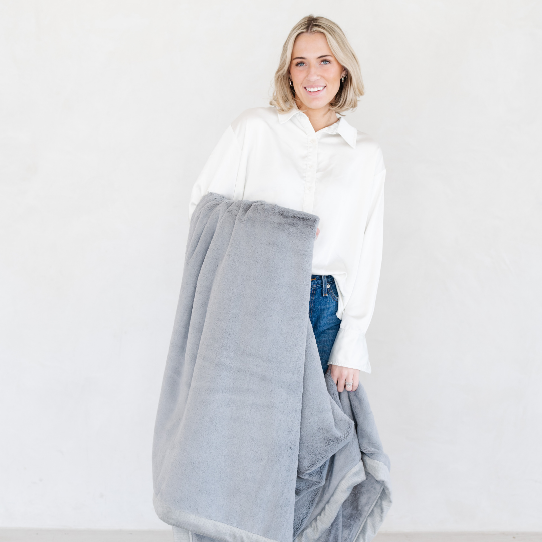 A woman smiles while holding a Gray Lush XL Saranoni blanket, designed for oversized comfort and featuring a buttery soft, plush texture. This luxurious Saranoni blanket is perfect for cozy evenings, reading, or home decor. The Saranoni blanket adds warmth, style, and unmatched softness, making it an ideal gift or essential for ultimate relaxation.