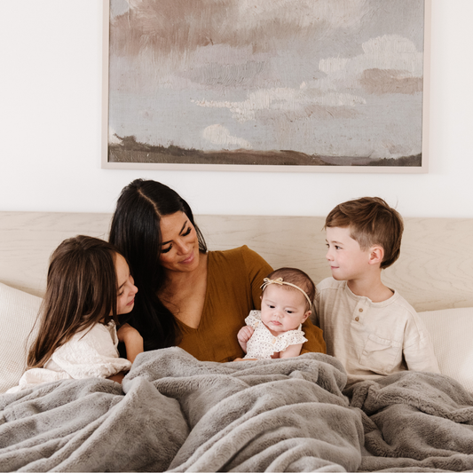 A mother and her children sit with a Saranoni Grand Faux Fur blanket, featuring an elegant gray tone and luxuriously soft texture. This cozy, queen-sized blanket and faux fur blanket offers soothing weight, premium softness, timeless comfort, and is a soft blanket. Perfect as a big blanket and luxury blanket for snuggling or as a luxury bedroom accent, this Saranoni blanket elevates every cozy moment and is a cozy blanket.