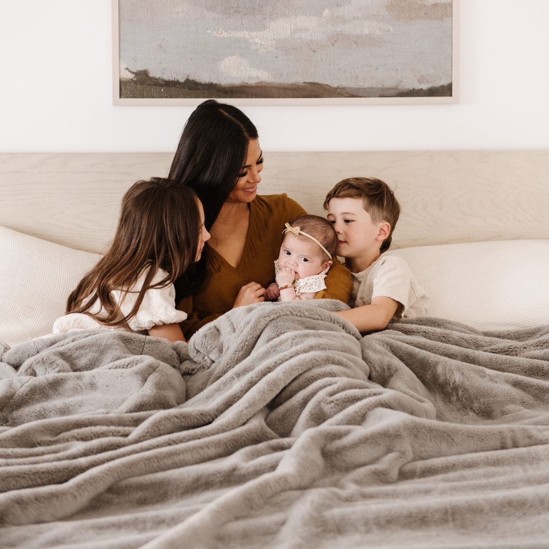 A mother and her two daughters and son share warm moments under a Saranoni Grand Faux Fur blanket, featuring an elegant gray tone and luxuriously soft texture. This cozy, queen-sized blanket and faux fur blanket offers soothing weight, premium softness, timeless comfort, and is a soft blanket. Perfect as a big blanket and luxury blanket for snuggling or as a luxury bedroom accent, this Saranoni blanket elevates every cozy moment and is a cozy blanket.