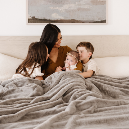 A mother and her two daughters and son share warm moments under a Saranoni Grand Faux Fur blanket, featuring an elegant gray tone and luxuriously soft texture. This cozy, queen-sized blanket and faux fur blanket offers soothing weight, premium softness, timeless comfort, and is a soft blanket. Perfect as a big blanket and luxury blanket for snuggling or as a luxury bedroom accent, this Saranoni blanket elevates every cozy moment and is a cozy blanket.