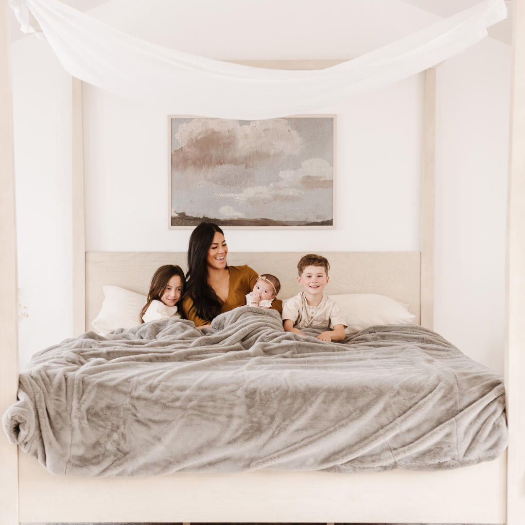 A mother and her children sit on a bed with a Saranoni Grand Faux Fur blanket, featuring an elegant gray tone and luxuriously soft texture. This cozy, queen-sized blanket and faux fur blanket offers soothing weight, premium softness, timeless comfort, and is a soft blanket. Perfect as a big blanket and luxury blanket for snuggling or as a luxury bedroom accent, this Saranoni blanket elevates every cozy moment and is a cozy blanket.