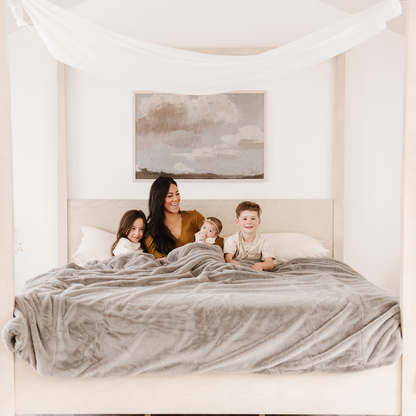A mother and her children sit on a bed with a Saranoni Grand Faux Fur blanket, featuring an elegant gray tone and luxuriously soft texture. This cozy, queen-sized blanket and faux fur blanket offers soothing weight, premium softness, timeless comfort, and is a soft blanket. Perfect as a big blanket and luxury blanket for snuggling or as a luxury bedroom accent, this Saranoni blanket elevates every cozy moment and is a cozy blanket.