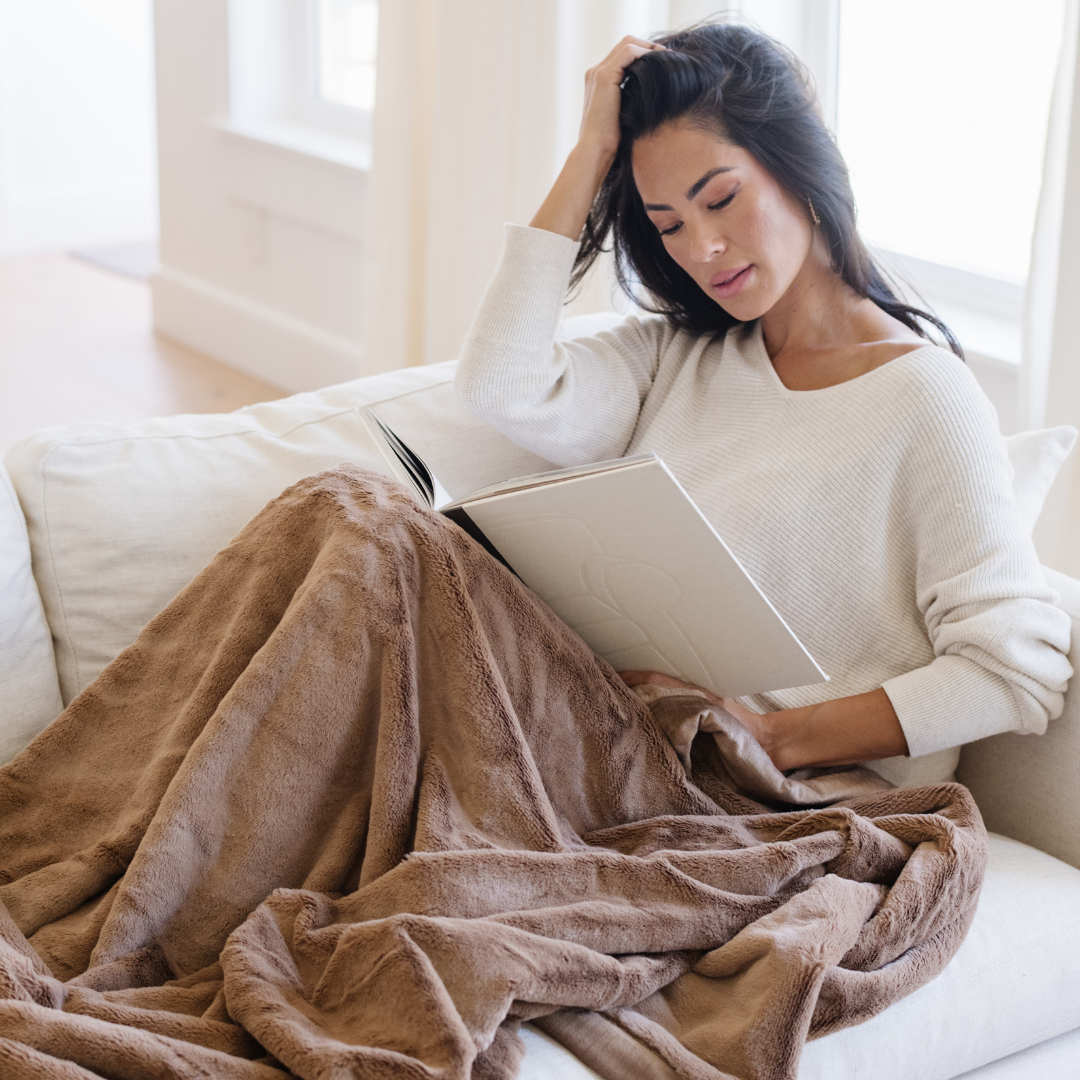 A woman reading on a white couch with a Hazelnut Lush XL Saranoni blanket, designed for oversized comfort and featuring a buttery soft, plush texture. This luxurious Saranoni blanket is perfect for cozy evenings, reading, or home decor. The Saranoni blanket adds warmth, style, and unmatched softness, making it an ideal gift or essential for ultimate relaxation.