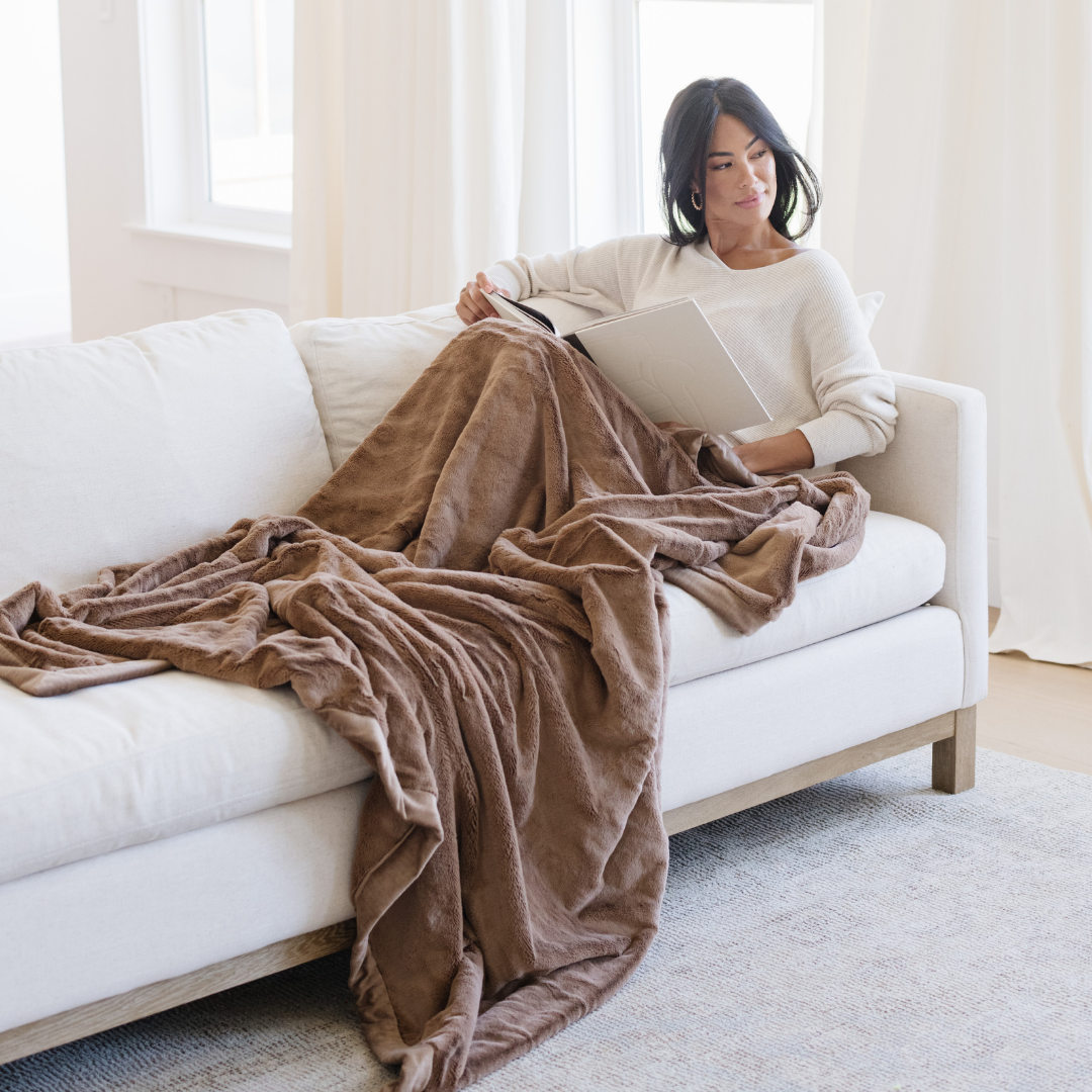 A woman relaxing on a white couch with a Hazelnut Lush XL Saranoni blanket, designed for oversized comfort and featuring a buttery soft, plush texture. This luxurious Saranoni blanket is perfect for cozy evenings, reading, or home decor. The Saranoni blanket adds warmth, style, and unmatched softness, making it an ideal gift or essential for ultimate relaxation.