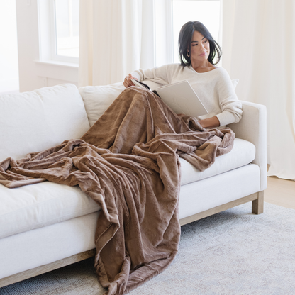 A woman relaxing on a white couch with a Hazelnut Lush XL Saranoni blanket, designed for oversized comfort and featuring a buttery soft, plush texture. This luxurious Saranoni blanket is perfect for cozy evenings, reading, or home decor. The Saranoni blanket adds warmth, style, and unmatched softness, making it an ideal gift or essential for ultimate relaxation.