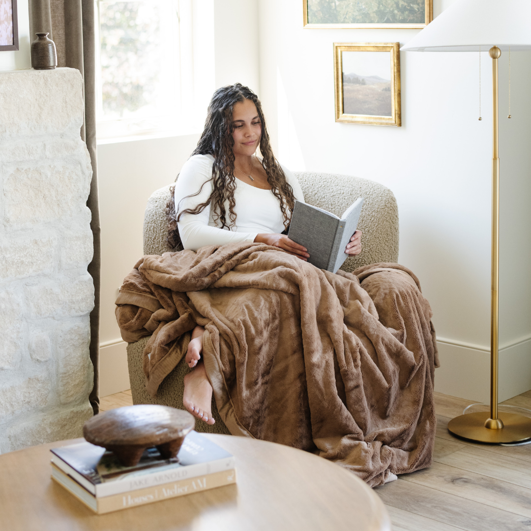 A woman reads a book and on her lap has a Hazelnut Lush XL Saranoni blanket, designed for oversized comfort and featuring a buttery soft, plush texture. This luxurious Saranoni blanket is perfect for cozy evenings, reading, or home decor. The Saranoni blanket adds warmth, style, and unmatched softness, making it an ideal gift or essential for ultimate relaxation.