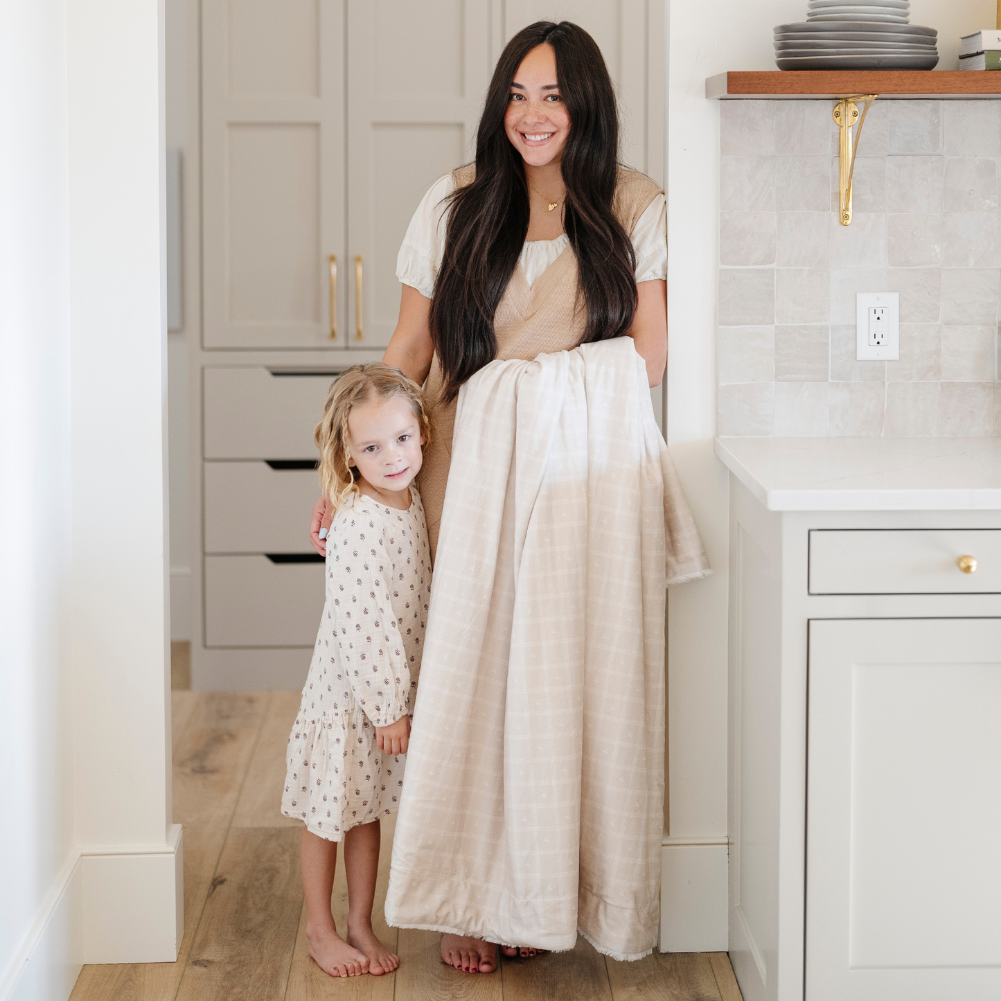 A mother stands by her little girl while holding a luxuriously soft Saranoni minky/lush blanket. This premium dual-textured extra-large throw features Saranoni’s signature cream lush fabric on one side and ultra-soft square printed minky on the other. Designed for ultimate relaxation, this plush blanket is perfect for snuggling, movie nights, and adding a touch of luxury to any home. This soft blanket is a luxury blanket and a Saranoni blanket. The cozy blanket is a comfortable blanket. 