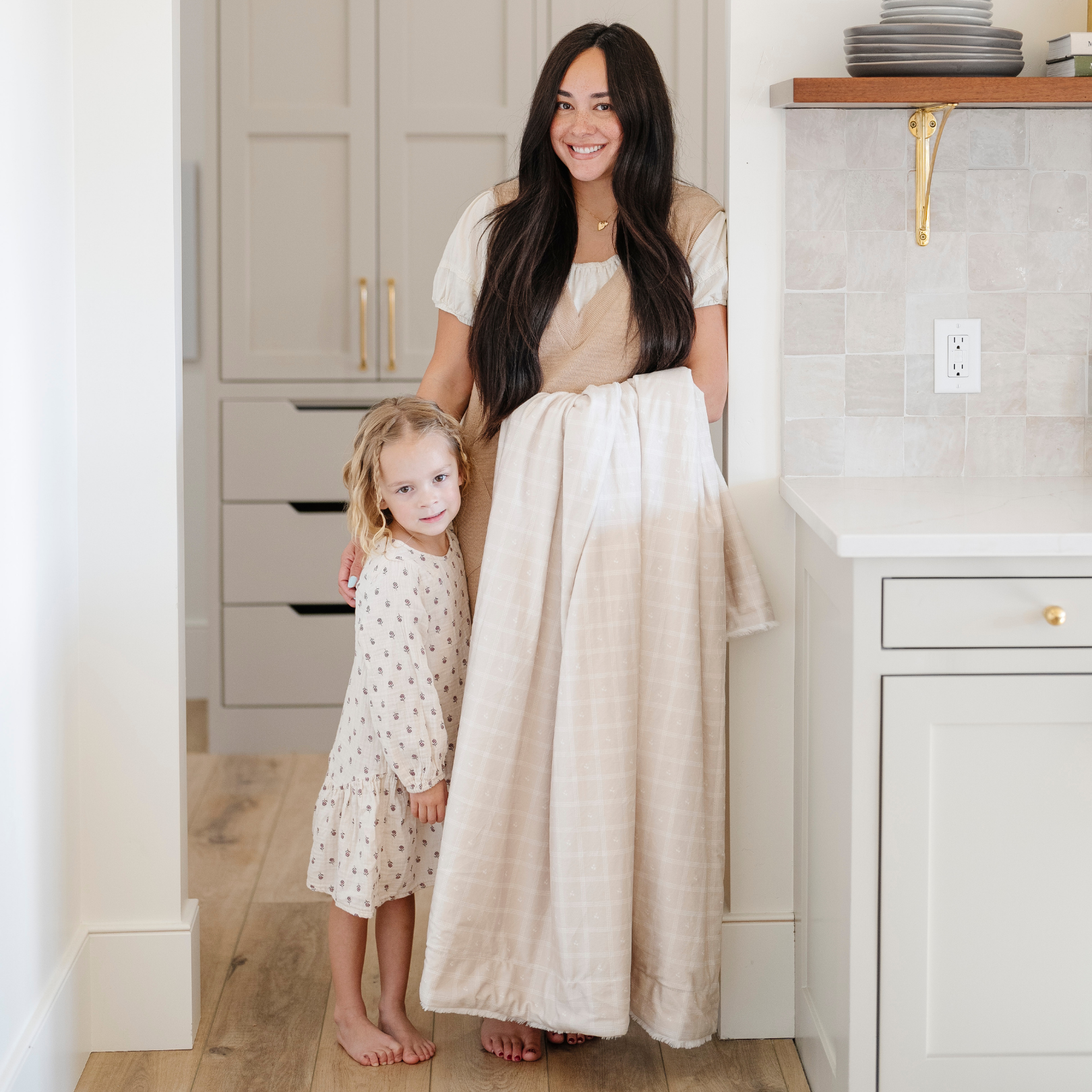 A mother stands by her little girl while holding a luxuriously soft Saranoni minky/lush blanket. This premium dual-textured extra-large throw features Saranoni’s signature cream lush fabric on one side and ultra-soft square printed minky on the other. Designed for ultimate relaxation, this plush blanket is perfect for snuggling, movie nights, and adding a touch of luxury to any home. This soft blanket is a luxury blanket and a Saranoni blanket. The cozy blanket is a comfortable blanket. 