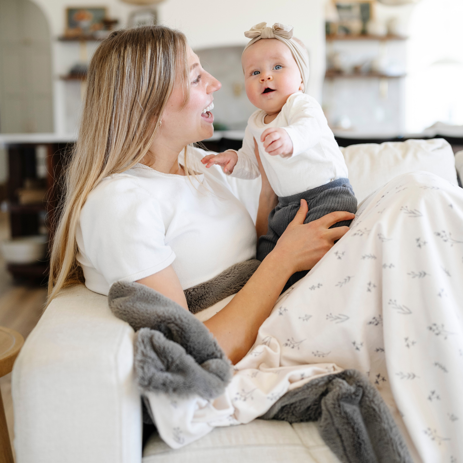 A mother sits on the couch holding her baby girl and sitting with a luxuriously soft Saranoni minky/lush blanket. This premium dual-textured throw blanket features Saranoni’s signature gray lush fabric on one side and ultra-soft floral printed minky on the other. This plush blanket is perfect for snuggling, movie nights, and adding a touch of luxury to any home. This soft blanket is a luxury blanket and a Saranoni blanket. The cozy blanket is a perfect comfort gift and is also beautiful for home decor. 