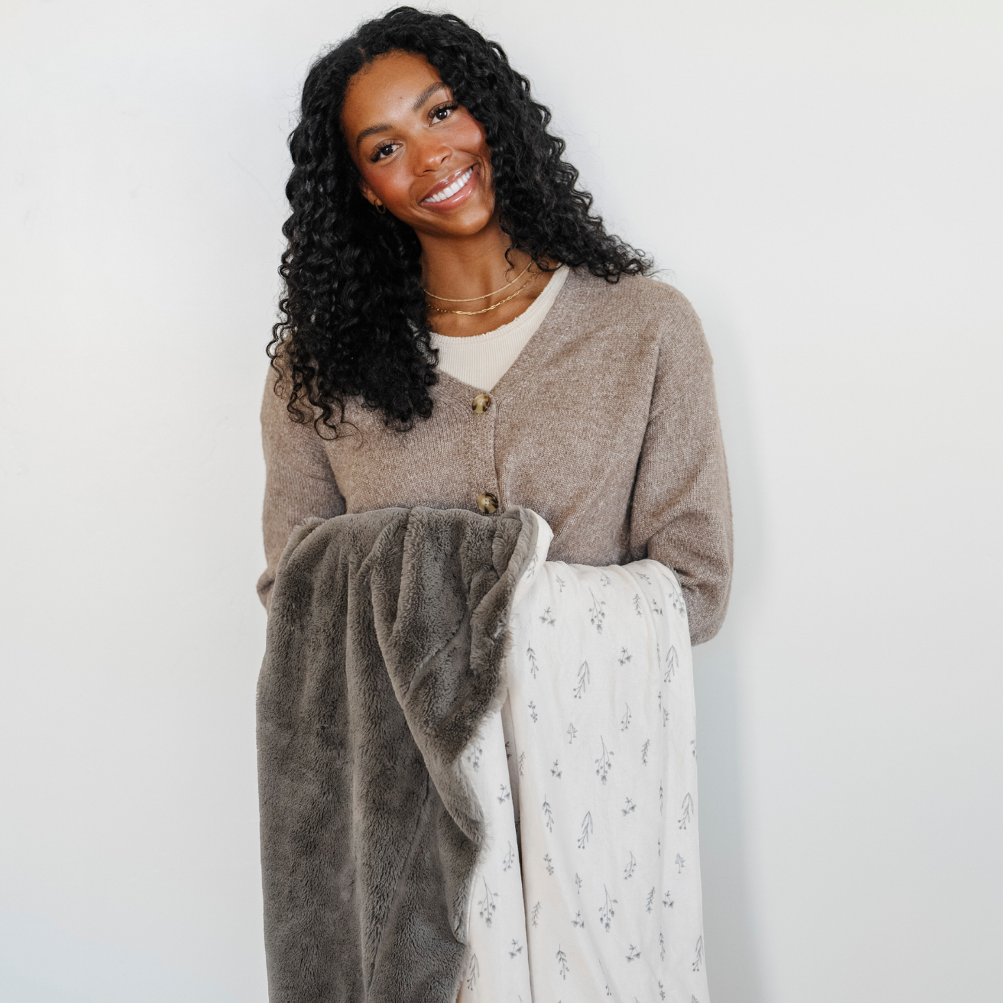 A woman smiles and holds a luxuriously soft Saranoni minky/lush blanket. This premium dual-textured extra-large throw blanket features Saranoni’s signature gray lush fabric on one side and ultra-soft floral printed minky on the other. This plush blanket is perfect for snuggling, movie nights, and adding a touch of luxury to any home. This soft blanket is a luxury blanket and a Saranoni blanket. The cozy blanket is a perfect comfort gift and is also beautiful for home decor. Enjoy this comfortable blanket!