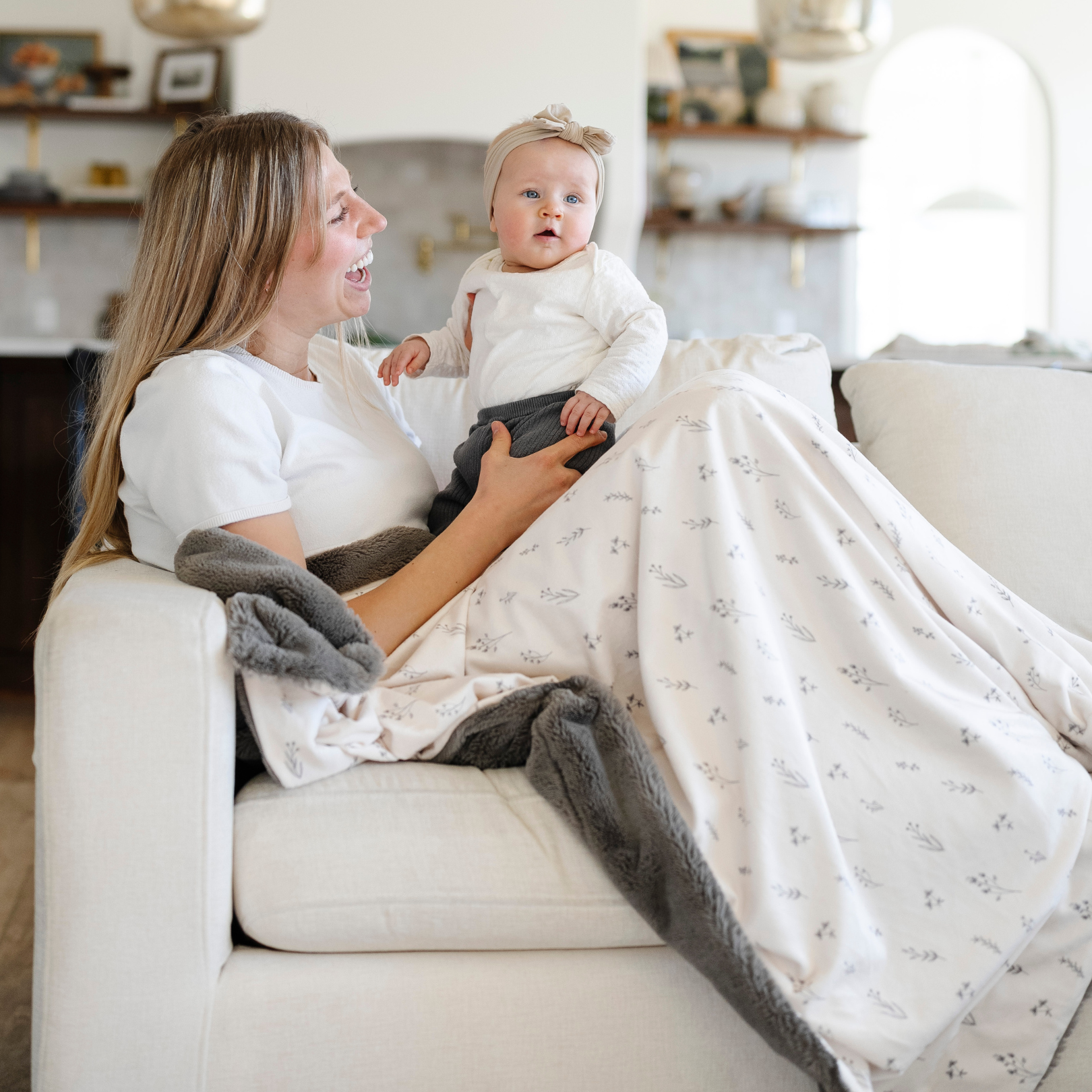A mother sits on the couch holding her baby girl and sitting with a luxuriously soft Saranoni minky/lush blanket. This premium dual-textured extra-large throw blanket features Saranoni’s signature gray lush fabric on one side and ultra-soft floral printed minky on the other. This plush blanket is perfect for snuggling, movie nights, and adding a touch of luxury to any home. This soft blanket is a luxury blanket and a Saranoni blanket. The cozy blanket is a perfect comfort gift.