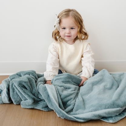 A little girl sits with a lush eucalyptus blanket. This Saranoni blanket is a toddler blanket and is a cute addition to a bedroom. This cozy blanket is also a great gift for toddlers. Enjoy this luxury blanket for years to come!