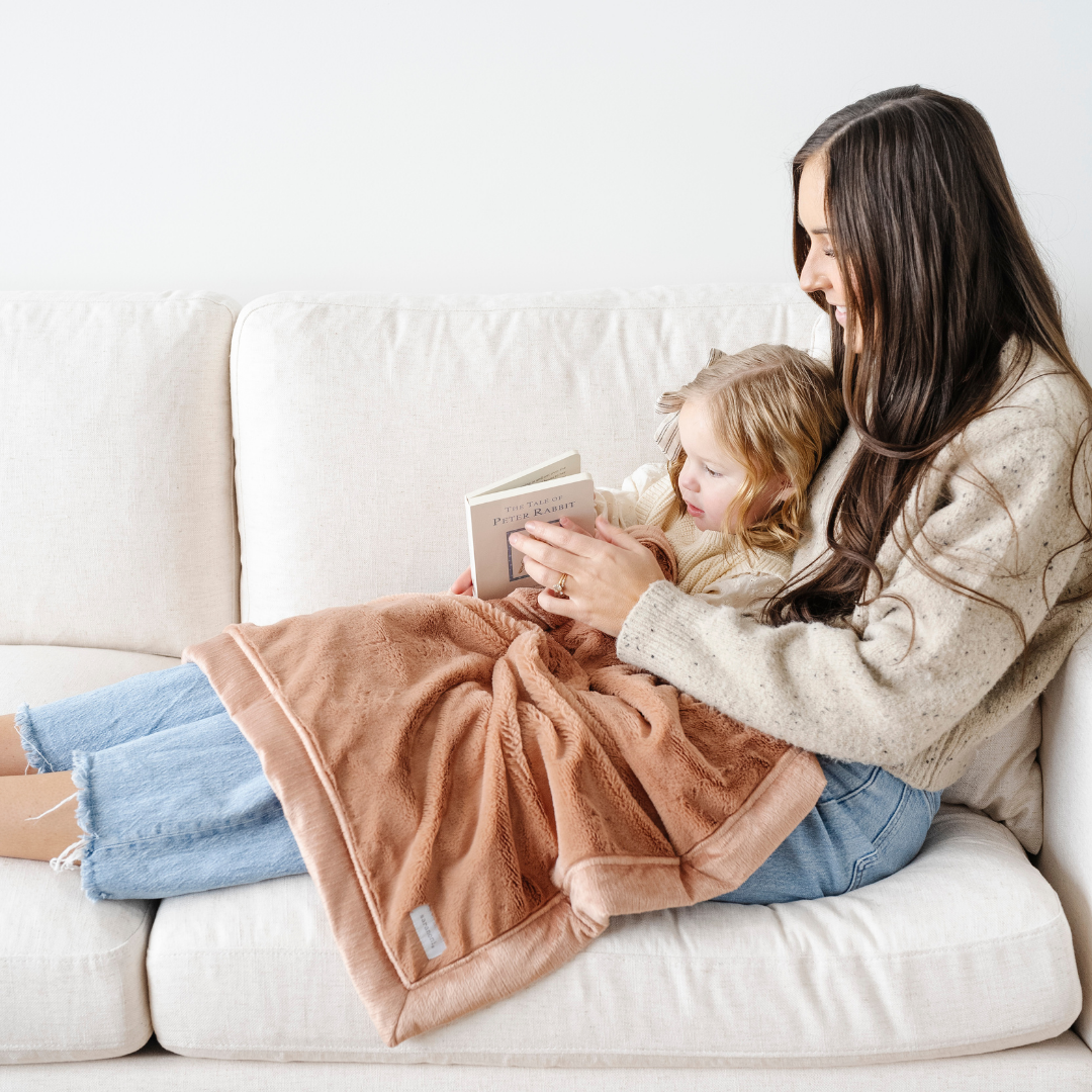 A mother and her daughter are sitting on a couch while reading. They have a peach lush blanket that is a receiving blanket on their laps. This soft blanket is a luxury blanket and a Saranoni blanket. This blanket is great for gifting or adding to a nursery or bedroom. 