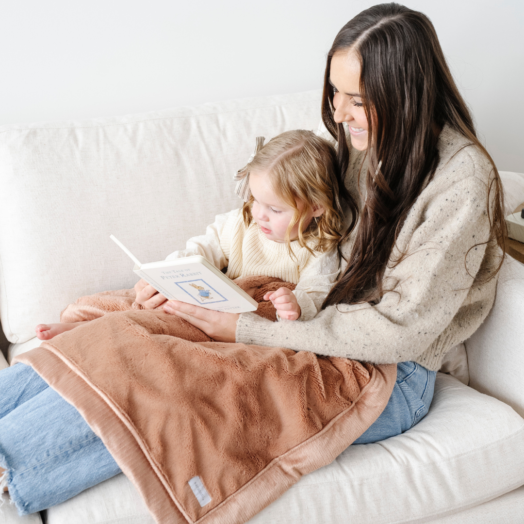 A mother is reading to her little girl. They have a peach colored lush blanket on their laps. This receiving blanket is a great size for littles and is a super soft blanket. Use this toddler blanket as a baby gift, kid's blanket, or an addition to a nursery. 