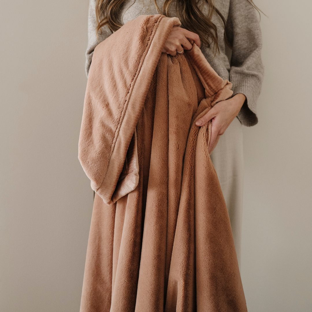 A woman holds a dark peach lush blanket that has a butter-soft texture. This soft blanket is a luxury blanket and a Saranoni blanket. This cozy blanket works great as a comfort gift and is also perfect for relaxing. 