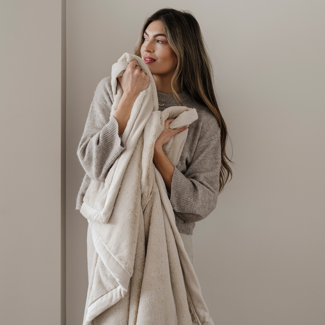 A woman holds a cream lush Saranoni blanket. This cozy blanket has a buttery-soft texture and is great for gifting or relaxing. You can also use this extra large blanket as a home decor staple. 