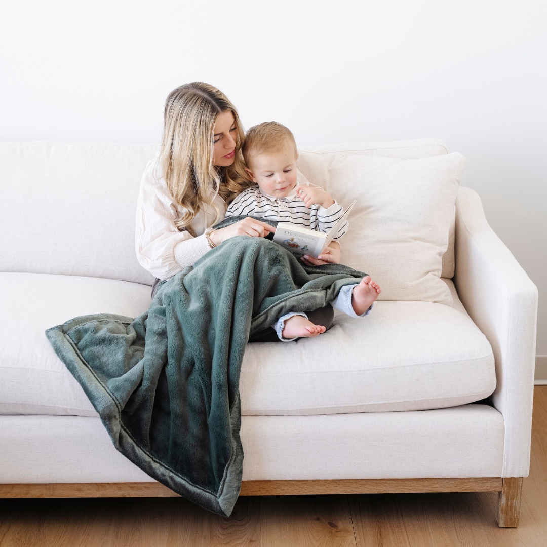 A mother and son sit on a couch. She is reading a book to him. They have a green Saranoni blanket on their laps. This soft blanket is a toddler blanket and is an adorable gift for toddlers. 