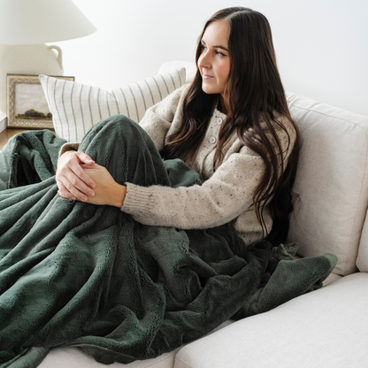 A woman sits on a couch with a green lush blanket on her lap. This soft blanket is a Saranoni blanket and a luxury blanket. This extra large blanket is a cozy comfort gift and is also great for relaxing. 