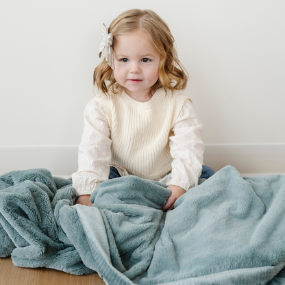 A little girl is sitting with a eucalyptus Saranoni blanket. This soft blanket is a receiving blanket and is a super cozy baby blanket. The most adorable addition to a nursery or bedroom, this lush blanket is made to last. 