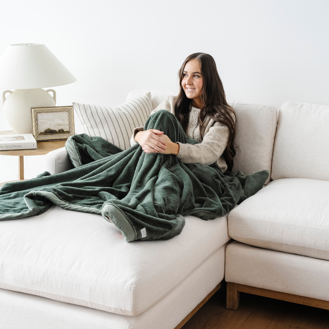 A smiling woman is sitting on a couch with a green blanket that is a lush blanket. This luxury blanket is great for relaxing, gifting, or as a home decor staple. This Saranoni blanket is also an extra large throw blanket. Enjoy this cozy blanket for years to come. 