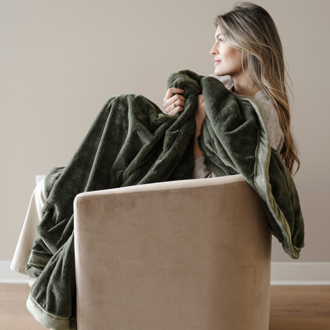A woman sitting in a chair is holding a green lush blanket. This luxury blanket is a soft blanket and a Saranoni blanket. This extra large throw blanket is great for gifting, relaxing, or as a home decor staple. Enjoy this cozy blanket for years to come. 